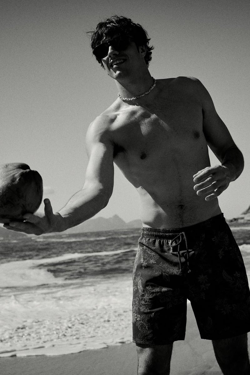 Francesco Ruggiero smiles in dark sunglasses while extending a coconut toward the camera, wearing a beaded necklace and dark hibiscus-print swim shorts on a wide sand beach, photographed in black and white