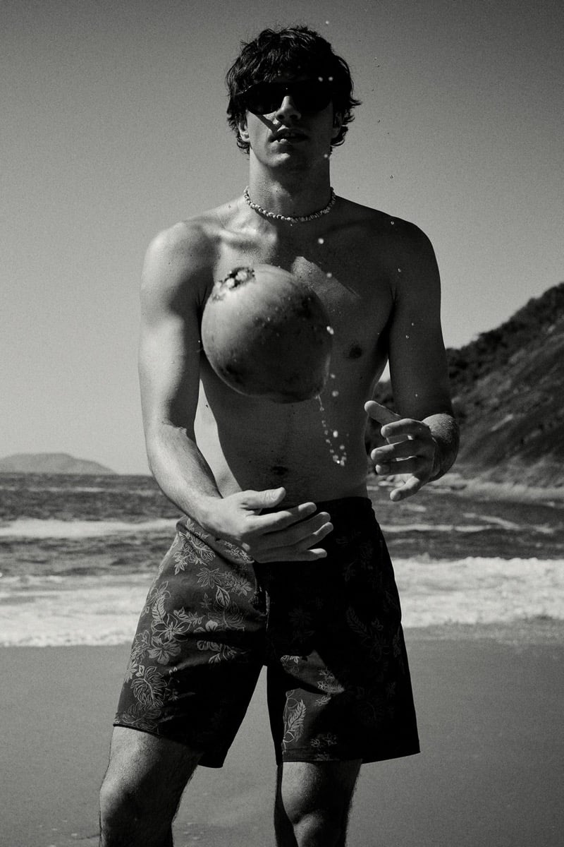 Francesco Ruggiero catches a falling coconut against his chest, water droplets in motion around him, wearing sunglasses, a beaded necklace, and dark hibiscus-print swim shorts, in black and white