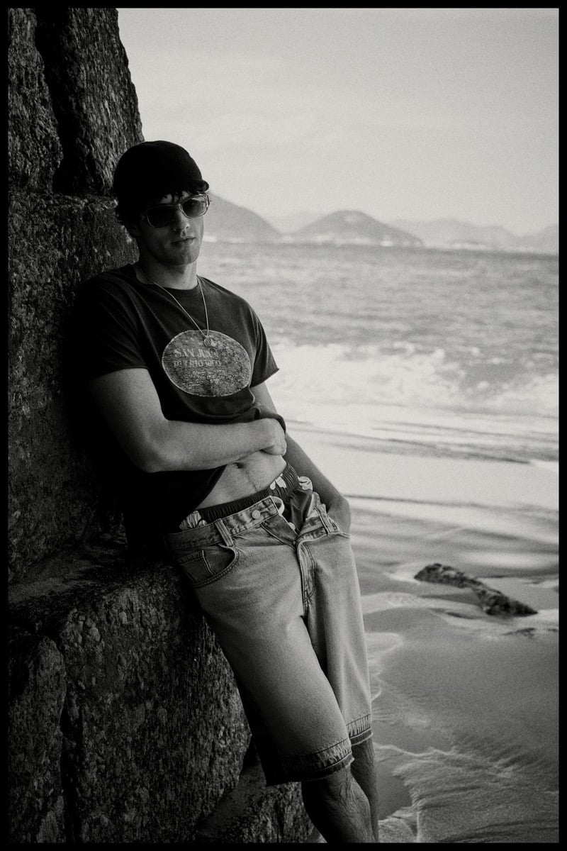 Francesco Ruggiero leans against a black volcanic rock wall in sunglasses, a backward baseball cap, a faded San Juan souvenir T-shirt, and washed denim shorts, photographed in black and white by the shore
