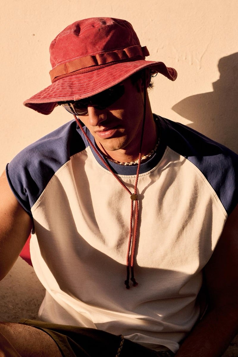 Francesco Ruggiero sits against a warm beige wall wearing a faded red wide-brim bucket hat with a cord, dark sunglasses, and a cream and navy raglan-sleeve tee with a beaded necklace