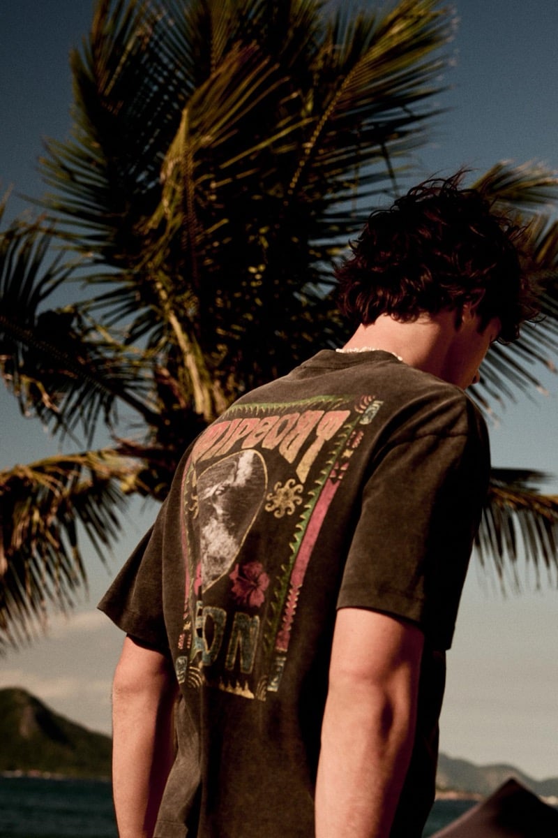Francesco Ruggiero stands with his back to the camera in front of a tall palm tree, wearing a washed dark graphic T-shirt with a tropical illustration printed across the back