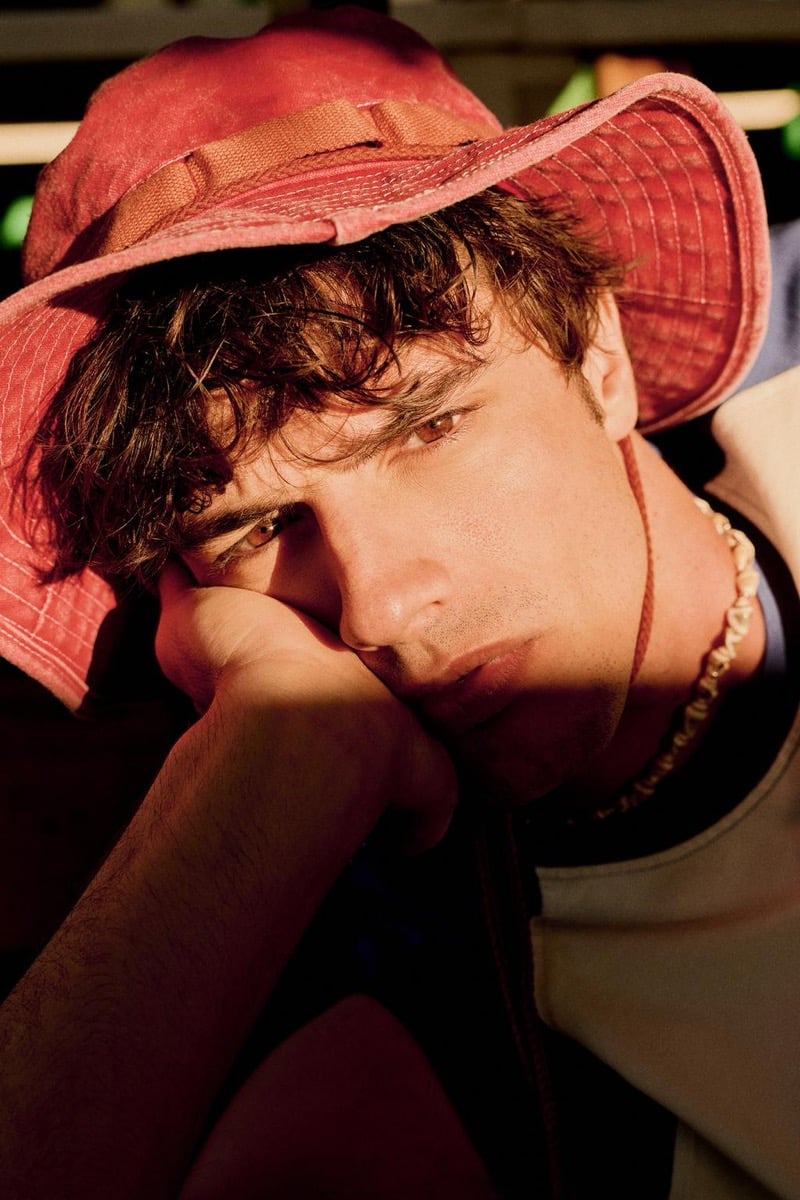 Intimate close-up of Francesco Ruggiero resting his head on his arm in a coral red corded bucket hat and a shell necklace, dappled warm light across his face