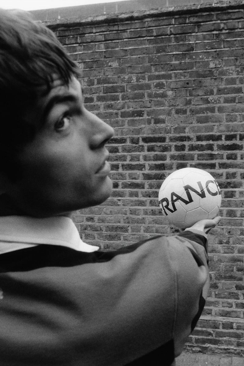 Black and white profile of Tom Meacock holding up a white soccer ball printed with the FRANCE branding from the 1998 World Cup, wearing a dark rugby shirt with a contrast white collar against a weathered brick wall