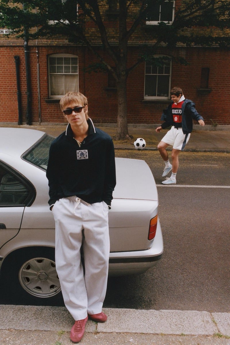 Joshua Boniface leaning back against a parked white sedan in a navy long-sleeve rugby polo with a FIFA Classics patch at the chest, cream pinstriped wide-leg trousers, pink suede trainers, and dark rectangular sunglasses, while Tom Meacock kicks a soccer ball in the background wearing a red England '66 jersey, navy track jacket, and white shorts