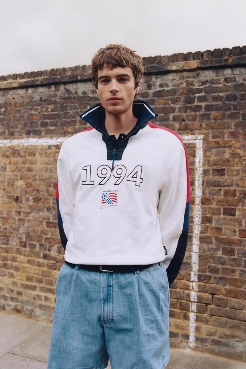 Tom Meacock standing against an outdoor brick wall in a white quarter-zip pullover with navy and red color-block panels down the sleeves, a '1994' graphic across the chest and the vintage USA '94 World Cup tag patch stitched below it, paired with light wash pleated denim shorts and a brown leather belt