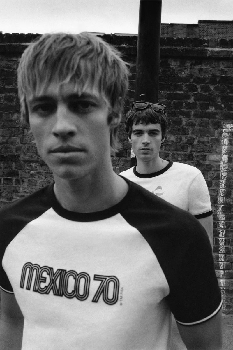 Black and white shot of Joshua Boniface in the foreground in a white raglan tee with black sleeves printed with the Mexico '70 logo and FIFA tag, and Tom Meacock standing behind him in a white ringer tee with black trim and sunglasses pushed up onto his forehead, against an exposed brick wall