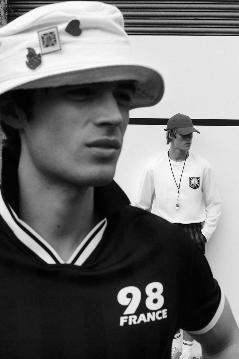 Black and white shot of Tom Meacock in the foreground wearing a white bucket hat trimmed with enamel pin badges and a black '98 France' polo with white tipping at the collar, while Joshua Boniface stands in the background in a red baseball cap, white long-sleeve crewneck with the England crest patch, and a silver whistle on a black cord