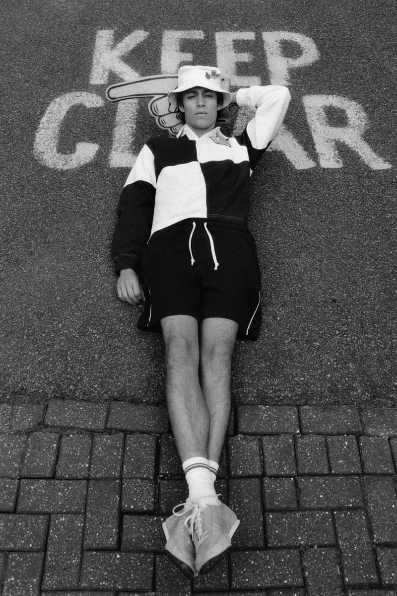 Joshua Boniface lying flat on gray asphalt painted with the words 'KEEP CLEAR' in white, wearing a black and white quartered rugby shirt, black drawstring shorts with white piping, a white bucket hat trimmed with enamel pin badges, white tube socks, and tan suede sneakers