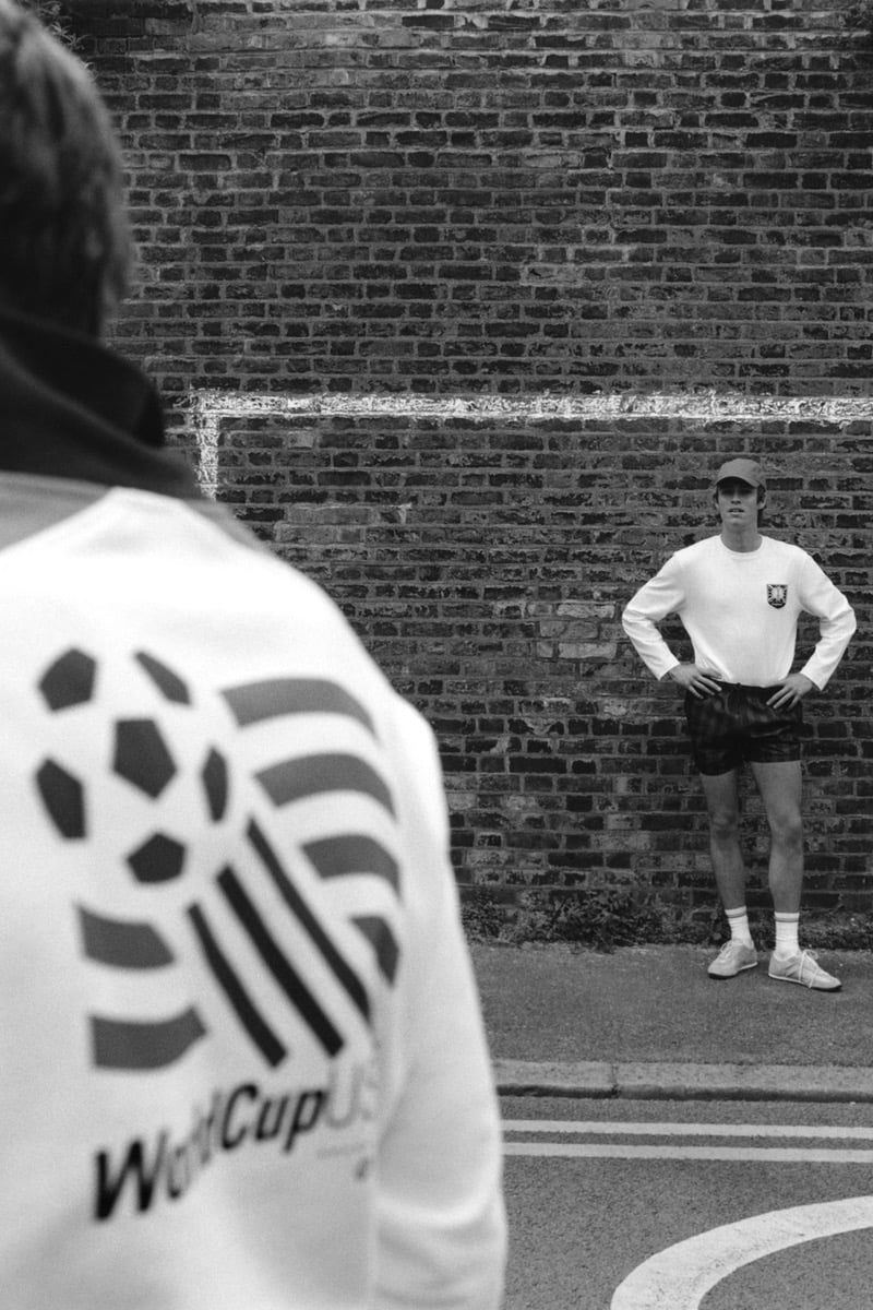 Black and white street shot of Tom Meacock seen from behind in a white long-sleeve tee printed with the WorldCup USA '94 stripes and soccer ball logo, with Joshua Boniface across the street leaning against a brick wall in a white long-sleeve with the England crest patch, dark patterned shorts, white tube socks, and a baseball cap