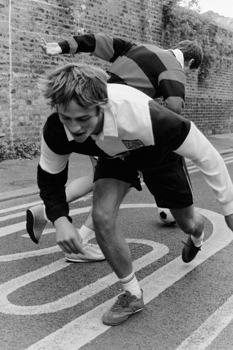 Black and white action shot of Joshua Boniface lunging forward across painted asphalt road markings in a black and white quartered rugby shirt, black drawstring shorts, white tube socks, and brown trainers, with Tom Meacock just behind in a navy and burgundy striped jersey and dark sneakers, a soccer ball between them
