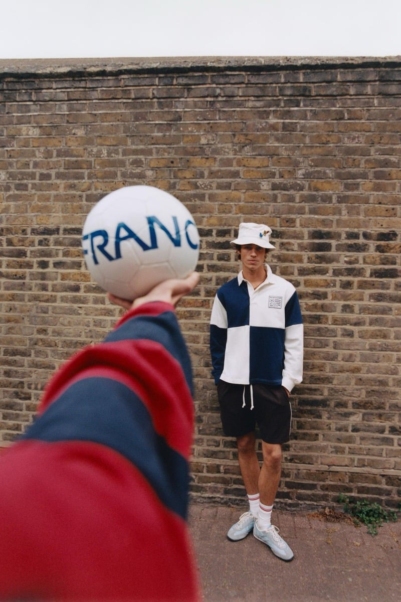 Joshua Boniface standing against a brick wall in a navy and white quartered long-sleeve rugby shirt with a FIFA Classics patch at the chest, black drawstring shorts, white tube socks, white sneakers, and a cream bucket hat, while a foreground hand in a red and navy striped sleeve extends a white France '98 soccer ball toward him