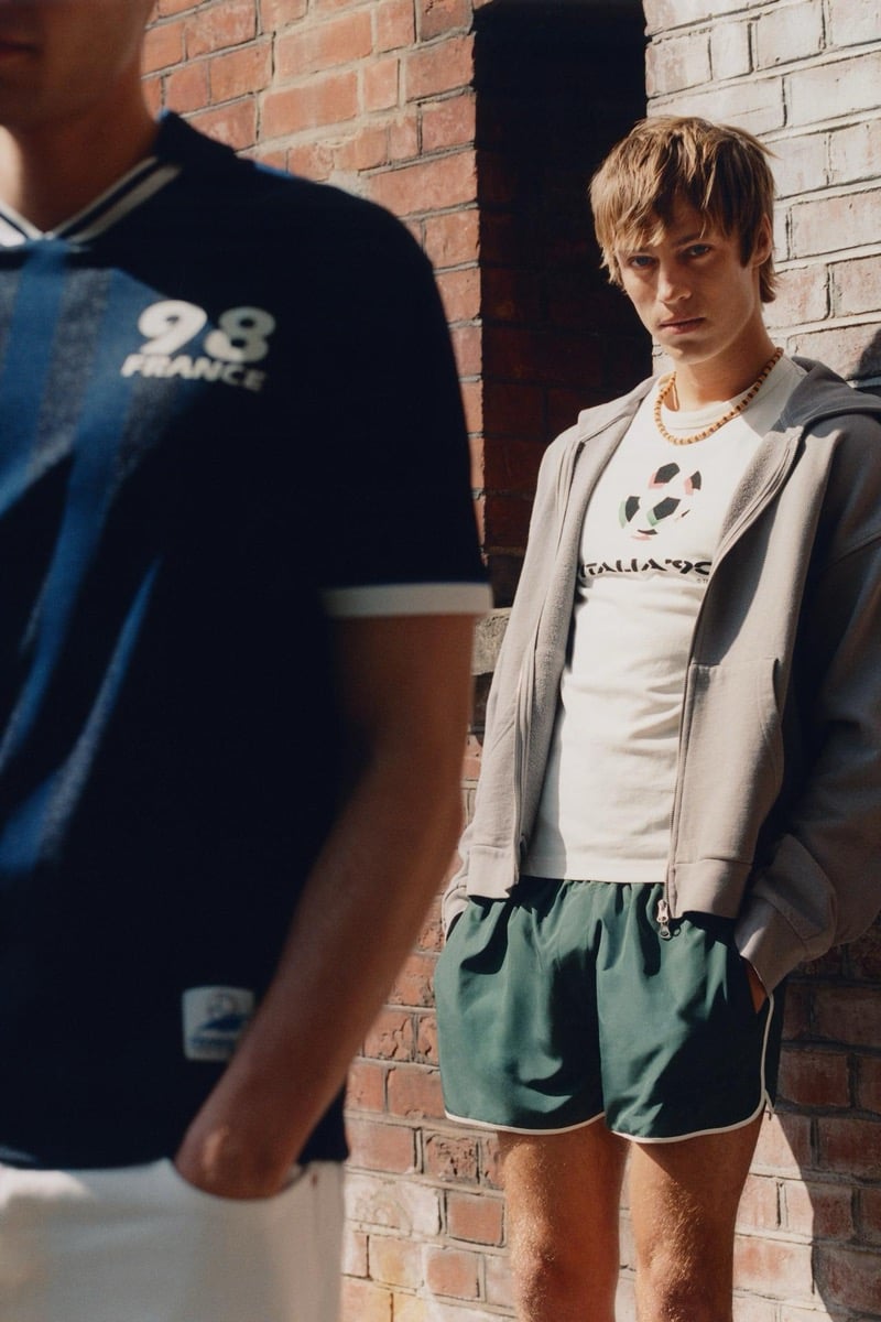 Joshua Boniface leaning against a brick wall in a white tee printed with the Italia '90 logo and a red and green soccer ball graphic, layered under an open gray zip-up hoodie, with forest green athletic shorts piped in white and a gold beaded necklace, while Tom Meacock stands out of focus in the foreground in a navy '98 France' jersey with the FIFA patch at the chest