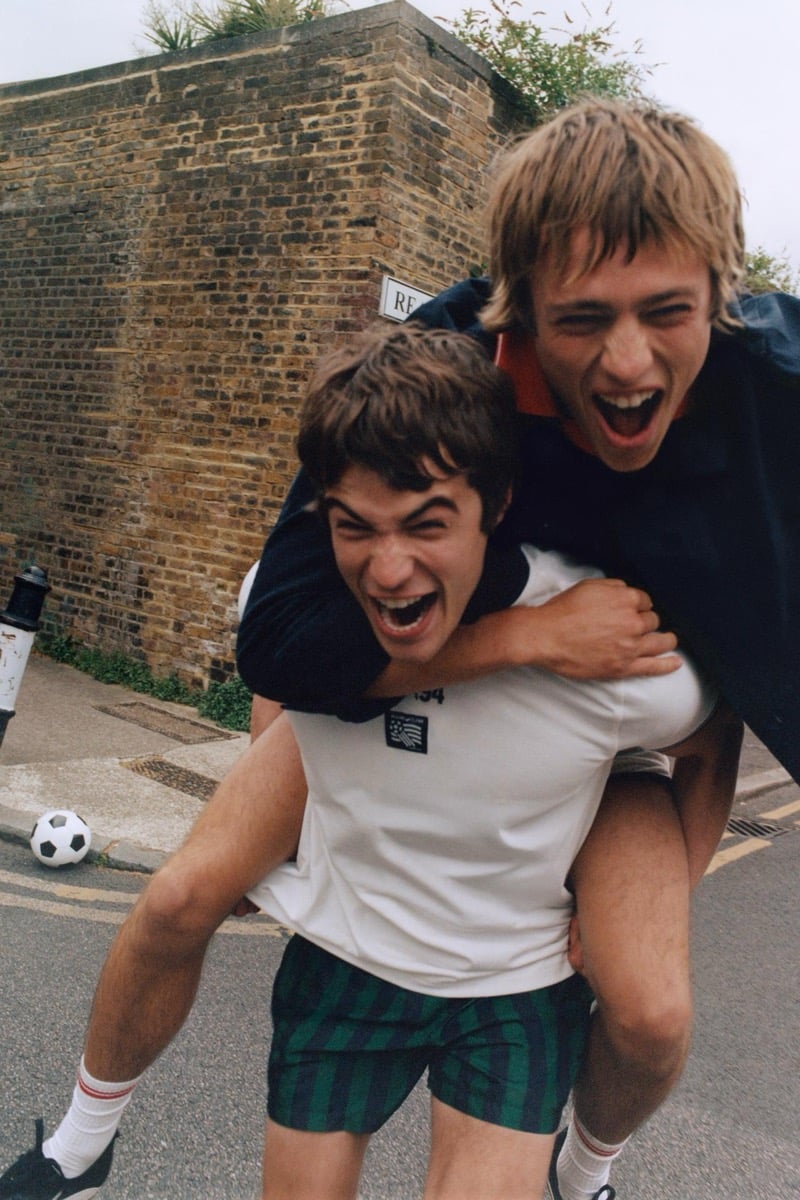 Joshua Boniface laughing on the back of Tom Meacock in a street piggyback, Tom in a white tee with the USA '94 tournament patch and green and navy plaid shorts, Joshua in a navy and red color-block polo and dark shorts, with a soccer ball at their feet