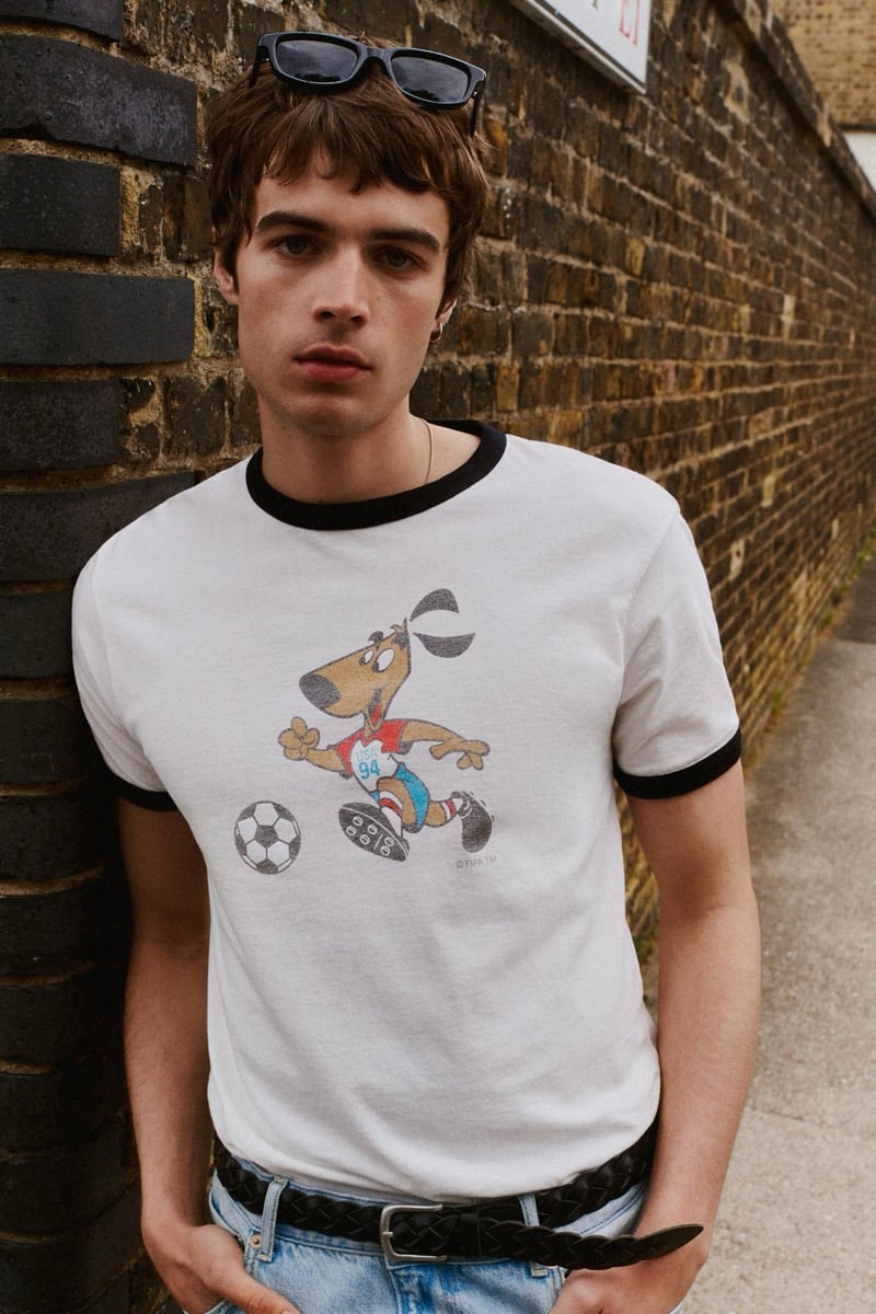 Tom Meacock leaning against an exposed brick wall in a white ringer tee trimmed in black at the sleeves and collar, printed with the cartoon Striker mascot from USA '94 dribbling a soccer ball, paired with light wash jeans, a braided black leather belt, and dark rectangular sunglasses pushed up onto his head