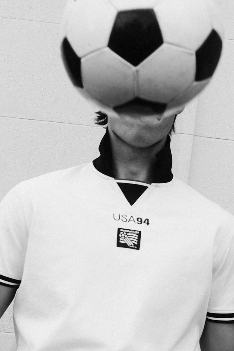 Black and white close shot of Tom Meacock with a black and white soccer ball pressed against his face, wearing a white polo printed with 'USA 94' and the embroidered World Cup logo patch