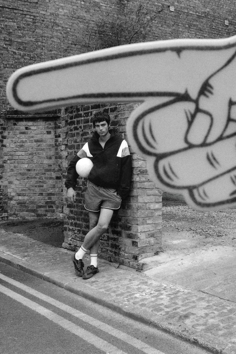 Black and white shot of Tom Meacock leaning against a brick archway in a navy half-zip pullover with white and red color-block shoulders, dark drawstring shorts with contrast piping, white tube socks, and brown trainers, holding a white soccer ball at his hip below an oversized graphic mural of a pointing hand