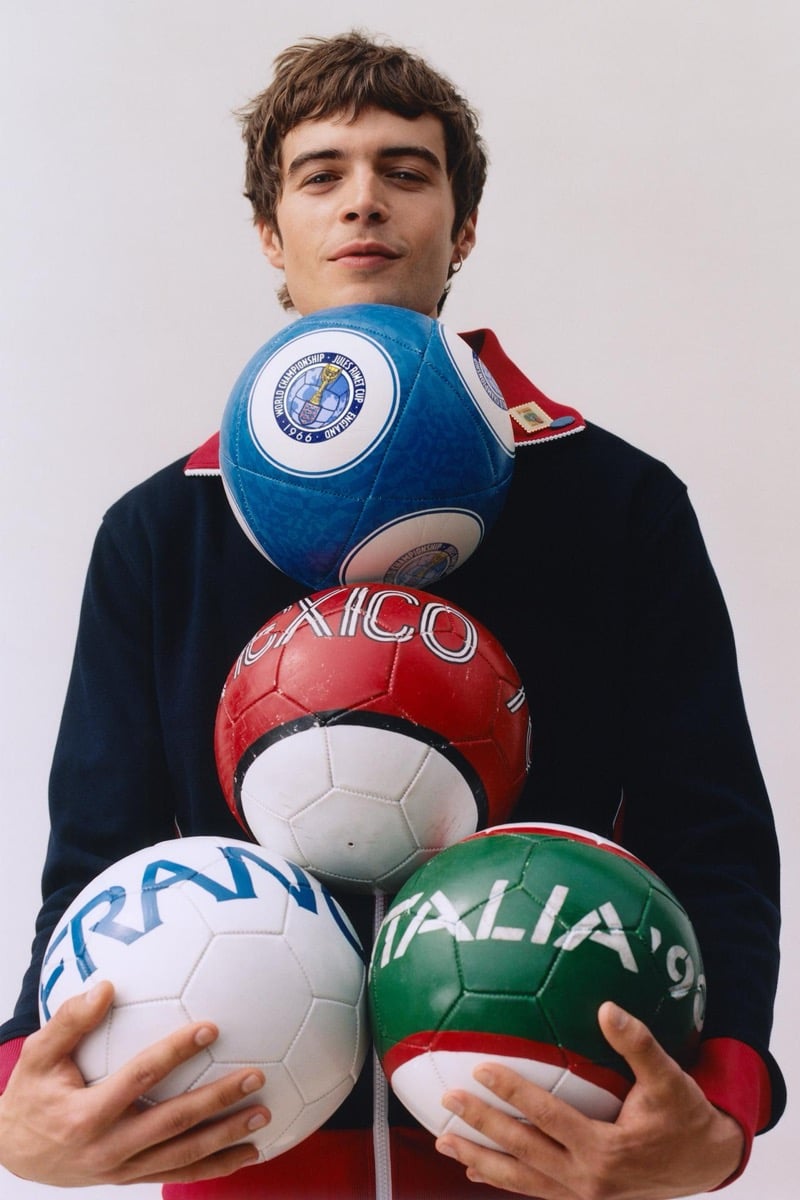 Tom Meacock stacking four vintage-style soccer balls against his chest — the blue England '66 commemorative ball, the red Mexico '70 ball, the white France '98 ball, and the green Italia '90 ball — wearing a navy and red panel-block quarter-zip rugby shirt against a soft studio backdrop