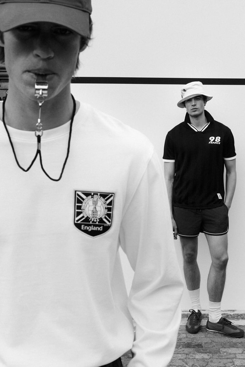 Black and white shot of Joshua Boniface in the foreground holding a silver whistle between his teeth on a black cord, wearing a dark baseball cap and a white long-sleeve crewneck with the England crest patch, while Tom Meacock stands in the background in a cream bucket hat, a black '98 France' polo with white tipping, dark drawstring shorts, white tube socks, and tan suede trainers