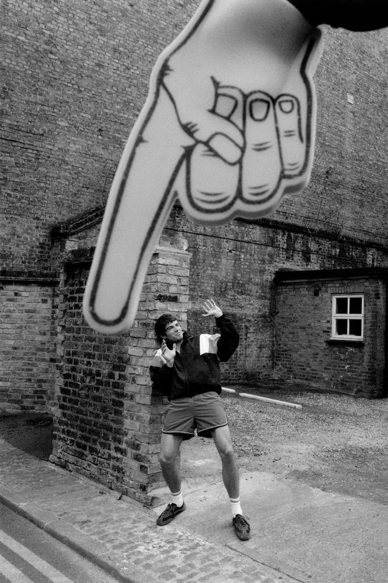 Black and white shot of Tom Meacock posed against a brick archway in a black collegiate-style varsity jacket with cream sleeves and contrast cuffs, dark drawstring shorts, white tube socks, and brown trainers, framed by an oversized graphic mural of a pointing hand
