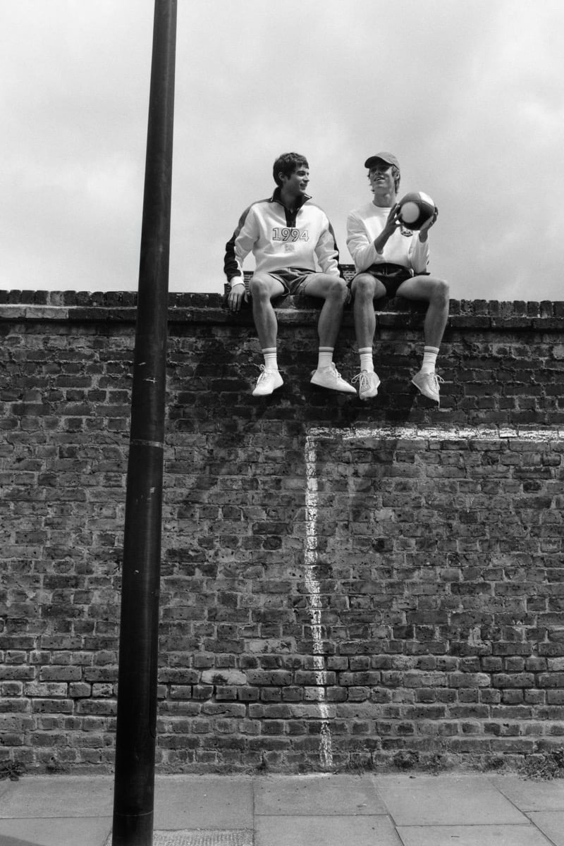 Black and white shot of Tom Meacock and Joshua Boniface sitting side by side on top of a brick wall above a chalk-drawn goal post, Tom in a white '1994' sweatshirt with navy raglan stripes, dark shorts, white socks, and white sneakers, Joshua in a white long-sleeve, dark shorts, baseball cap, and white sneakers, holding a soccer ball at his side