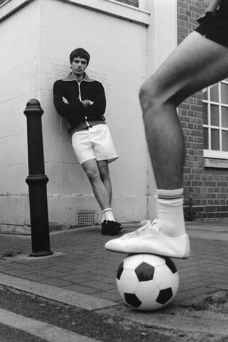 Black and white shot of Tom Meacock leaning against the corner of a stone building in a black zip-up track jacket with cream trim and white drawstring shorts, with Joshua Boniface's leg and foot in the foreground resting on a black and white soccer ball, in white sneakers and white tube socks