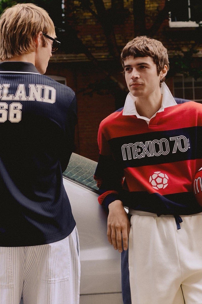 Joshua Boniface seen from behind in a navy long-sleeve England '66 polo paired with cream pinstriped trousers and dark rectangular sunglasses, alongside Tom Meacock in a red and navy striped Mexico '70 rugby shirt with a white soccer ball graphic and cream wide-leg trousers, both leaning against the white panel of a parked sedan on a London street lined with red brick