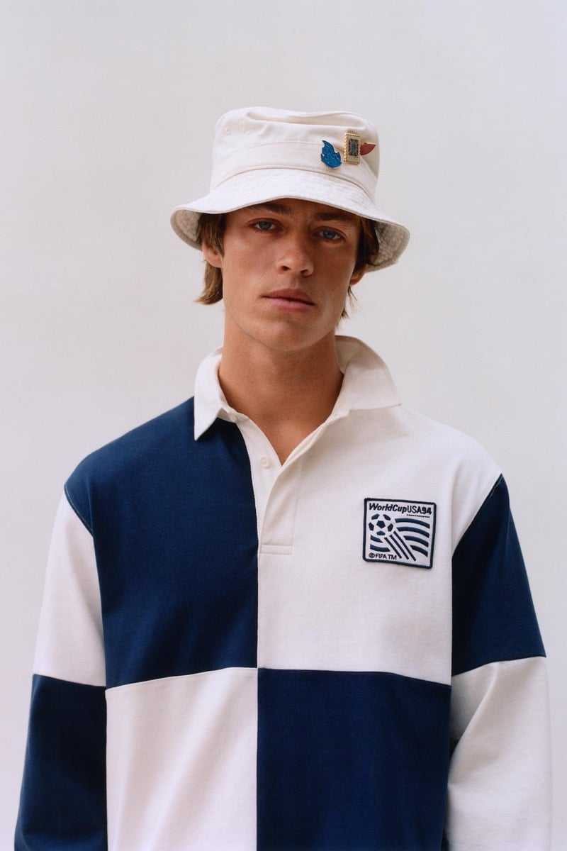 Joshua Boniface in a white bucket hat trimmed with enamel pin badges and a navy and white quartered long-sleeve rugby shirt with the WorldCup USA '94 FIFA patch at the chest, against a pale studio backdrop