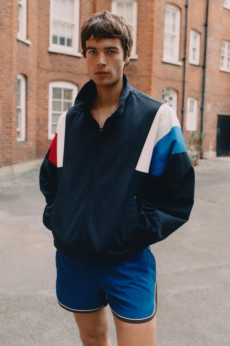 Tom Meacock standing in front of a red brick townhouse in a navy nylon zip-up windbreaker with white, red, and royal blue color-block panels across the shoulders and sleeves, paired with royal blue running shorts piped in white and black and a slim silver chain necklace