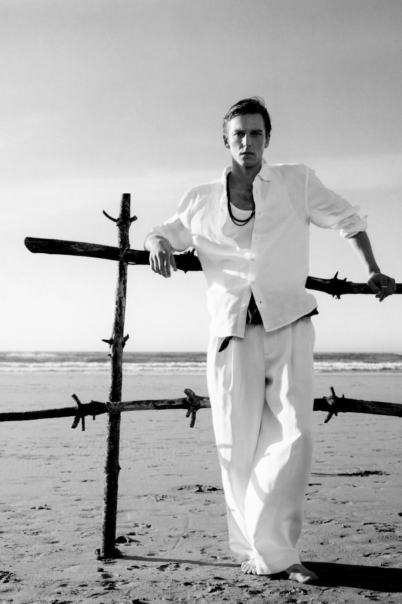 Quentin Demeester wears an open white linen shirt and wide-leg white trousers, leaning against a weathered driftwood cross on the beach in a black-and-white image