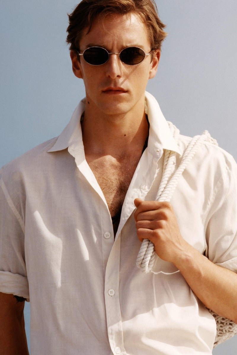 Close-up of Quentin Demeester in slim oval sunglasses and an unbuttoned white linen shirt, a woven rope-handled bag slung over his shoulder against a clear sky