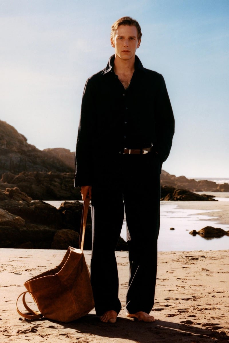 Quentin Demeester stands barefoot on a rocky beach in a dark long-sleeve shirt and loose trousers cinched with a brown leather belt, holding an oversized suede tote bag at his side