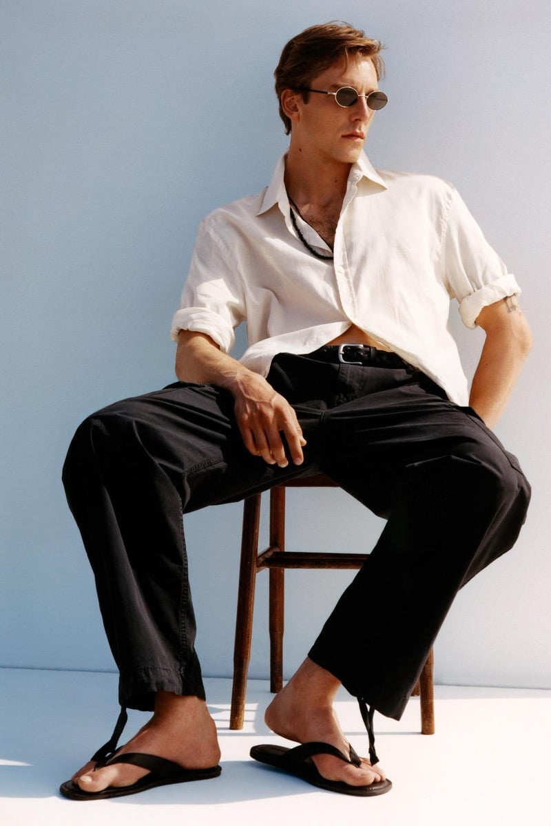 Quentin Demeester sits on a wooden stool in a half-buttoned white linen shirt, dark relaxed trousers, oval sunglasses, and leather thong sandals