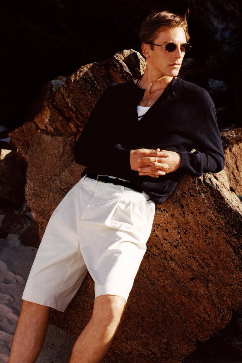 Quentin Demeester wears a dark V-neck knit over a white tank with pleated white shorts and slim oval sunglasses, leaning against rust-colored coastal rocks