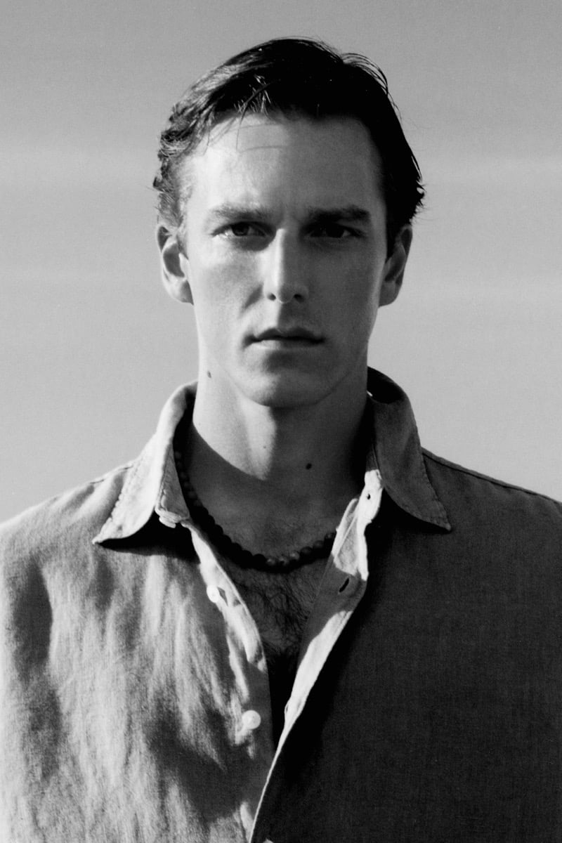 Black-and-white portrait of Quentin Demeester in an open camp-collar linen shirt with a beaded necklace, photographed against a soft sky