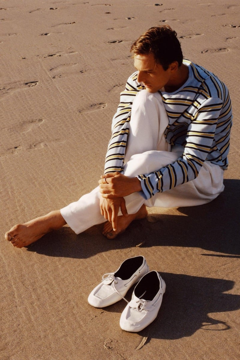 Quentin Demeester sits barefoot on the sand in a blue-striped long-sleeve top and loose white trousers, a pair of white boat shoes placed beside him
