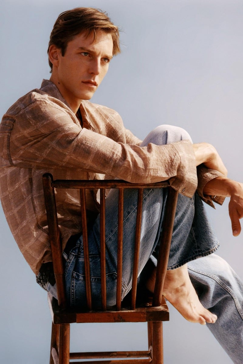 Quentin Demeester sits backward on a wooden chair in a tan windowpane-check linen overshirt and faded blue jeans, looking over his shoulder against a pale backdrop