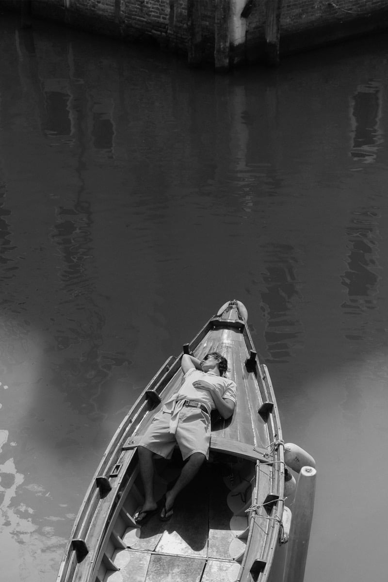 Black-and-white aerial image of model Tony Özkan stretched out in a wooden boat floating on a Venetian canal, wearing a short-sleeve shirt and cargo shorts