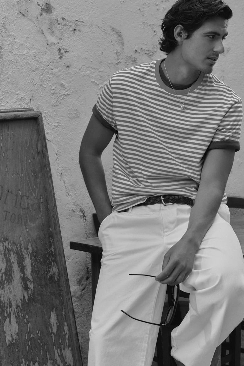 Black-and-white portrait of model Tony Özkan in a striped ringer tee tucked into light wide-leg trousers, holding sunglasses while seated against a stucco wall in Venice