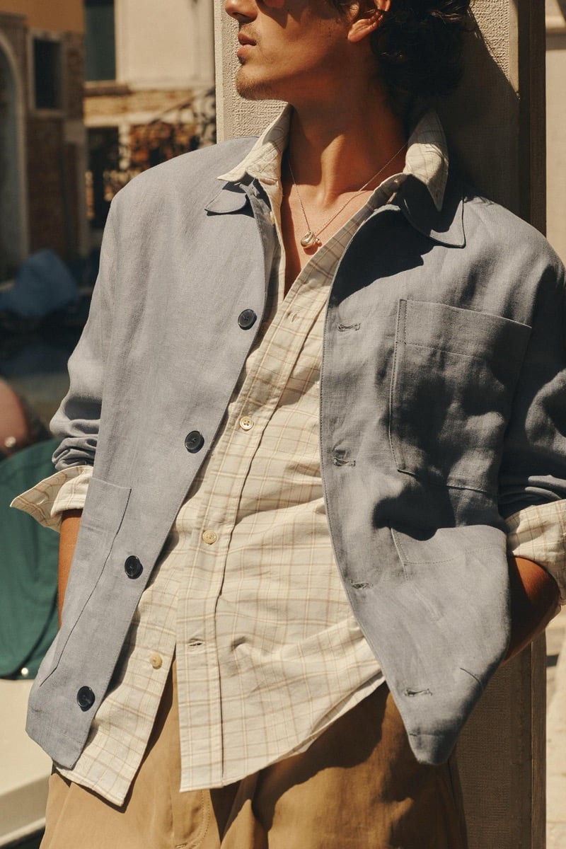 Sunlit close-up of model Tony Özkan in a faded blue linen overshirt worn over a tan and white checked shirt with light trousers, leaning into a shaded Venetian doorway
