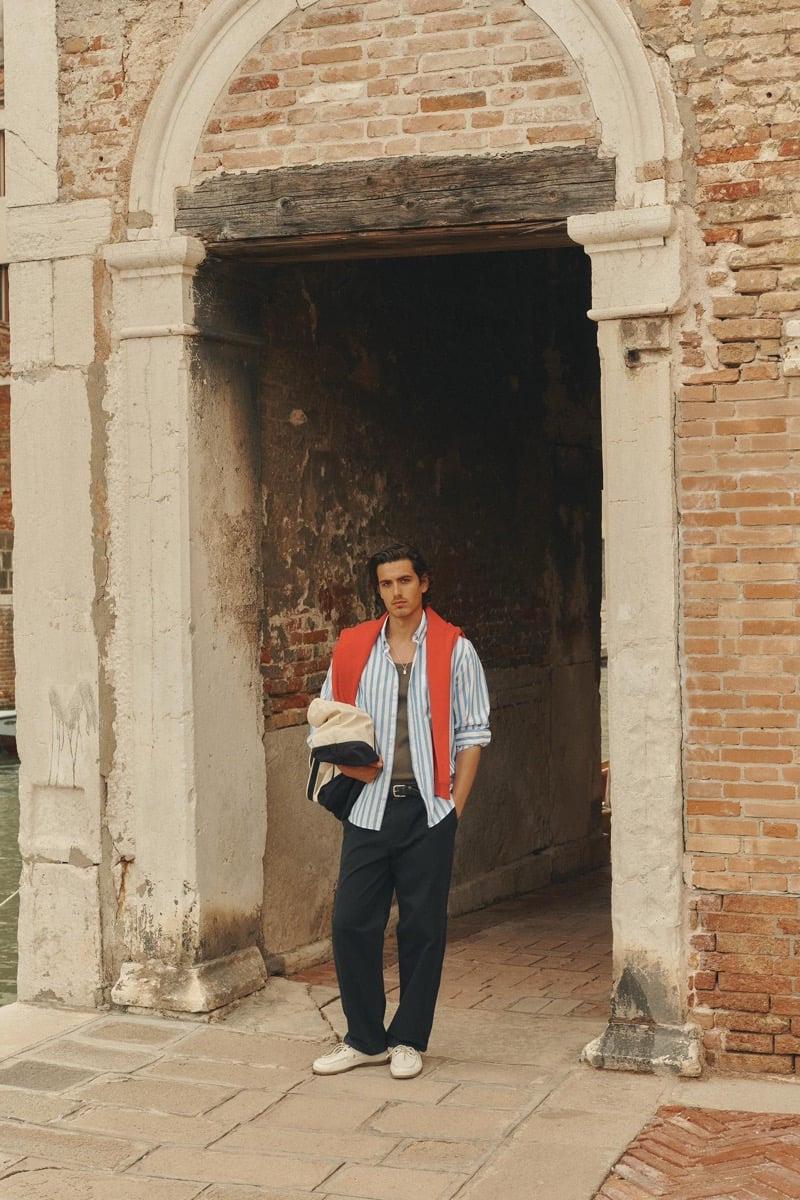Model Tony Özkan framed in a stone archway in Venice wearing a coral knit layered over a blue and white striped button-down with dark trousers and white sneakers