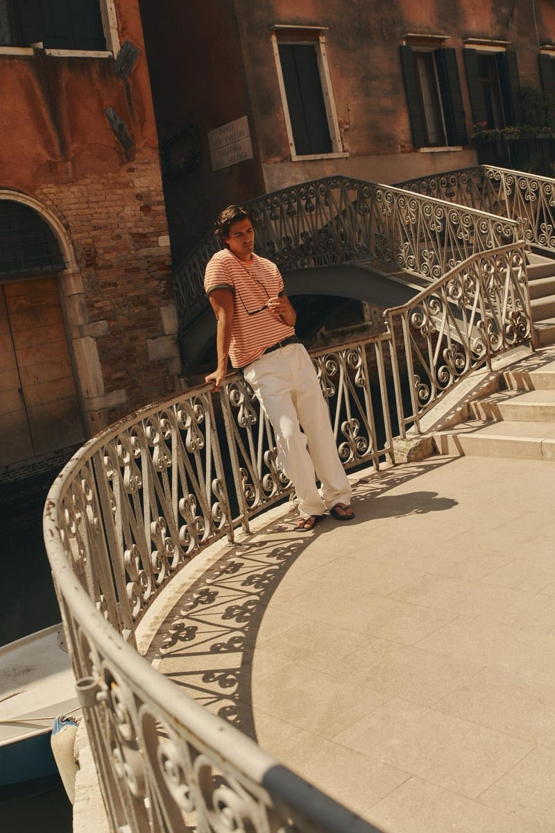 Model Tony Özkan leaning on the railing of a wrought-iron Venetian footbridge in an orange striped tee and light wide-leg trousers