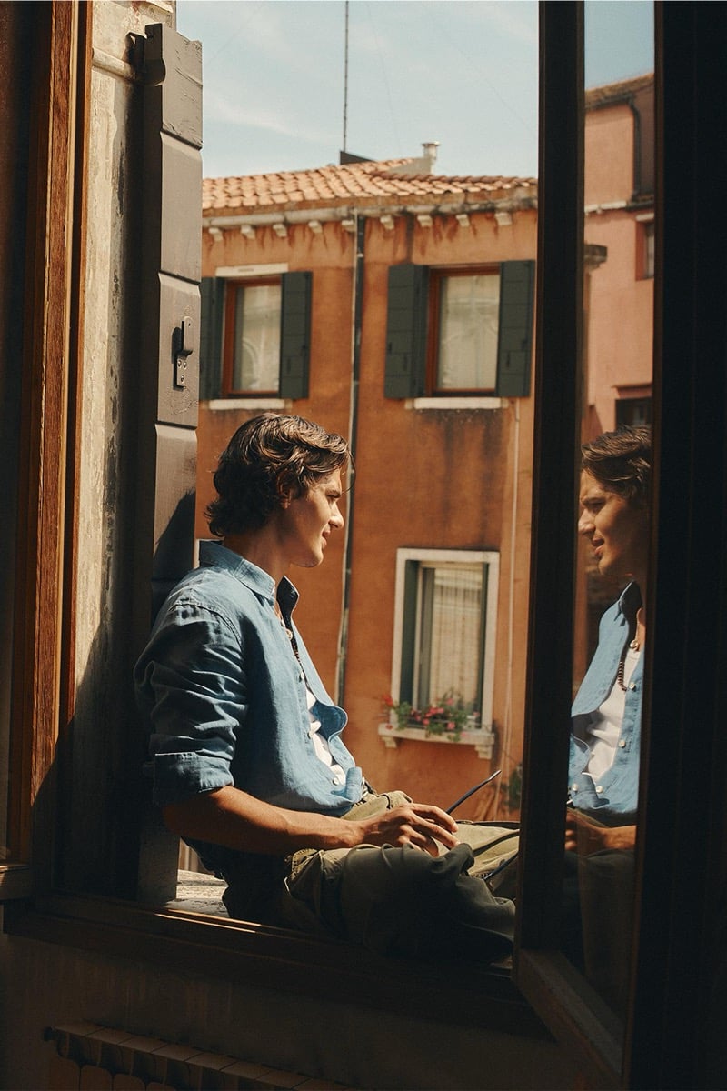 Model Tony Özkan seated on an open window ledge in a blue chambray shirt and olive shorts, framed by Venetian shutters with terracotta rooftops in the background
