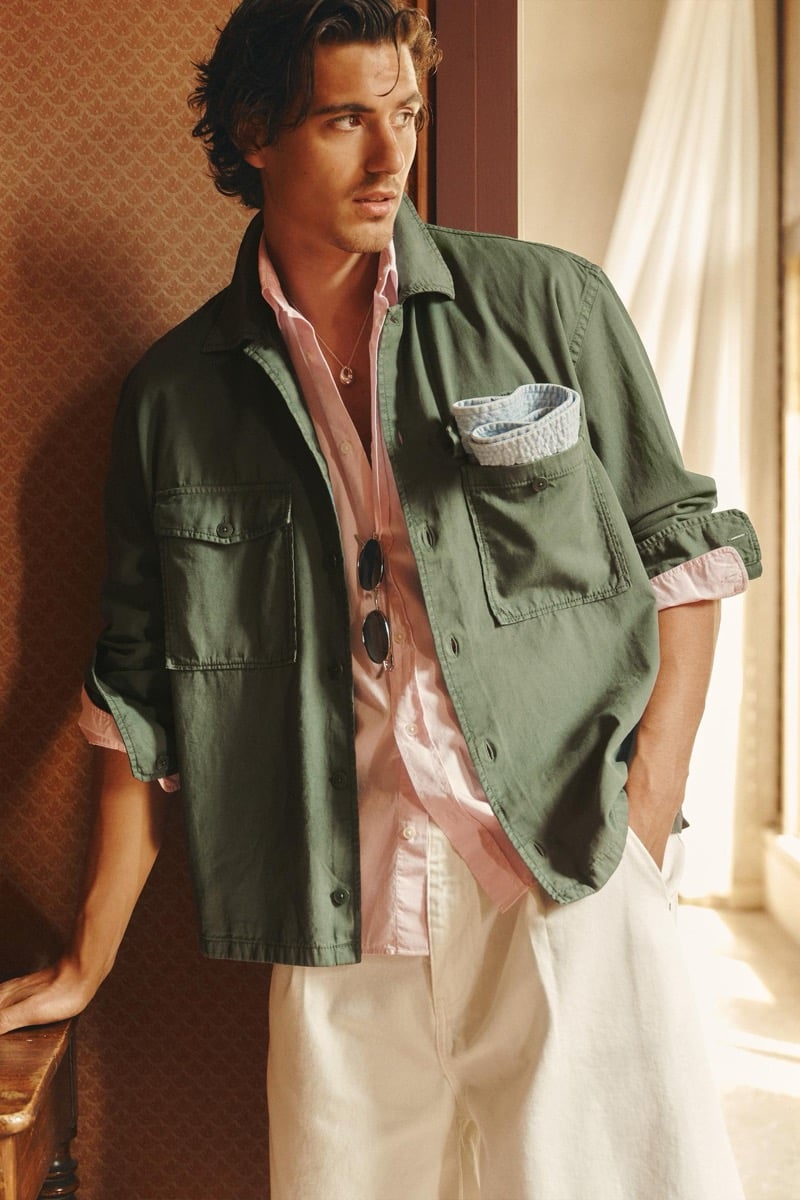 Model Tony Özkan in a sage green cotton overshirt layered over a pink button-down shirt and cream wide-leg pants, holding sunglasses against an interior wallpapered wall
