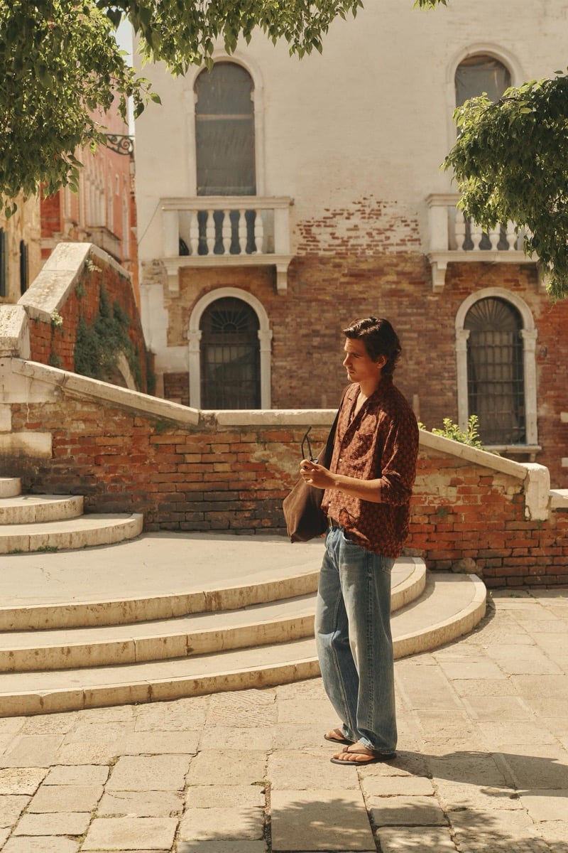 Model Tony Özkan walking through a Venetian piazza in a rust-toned printed shirt with faded blue jeans and brown sandals, framed by an arched stone facade