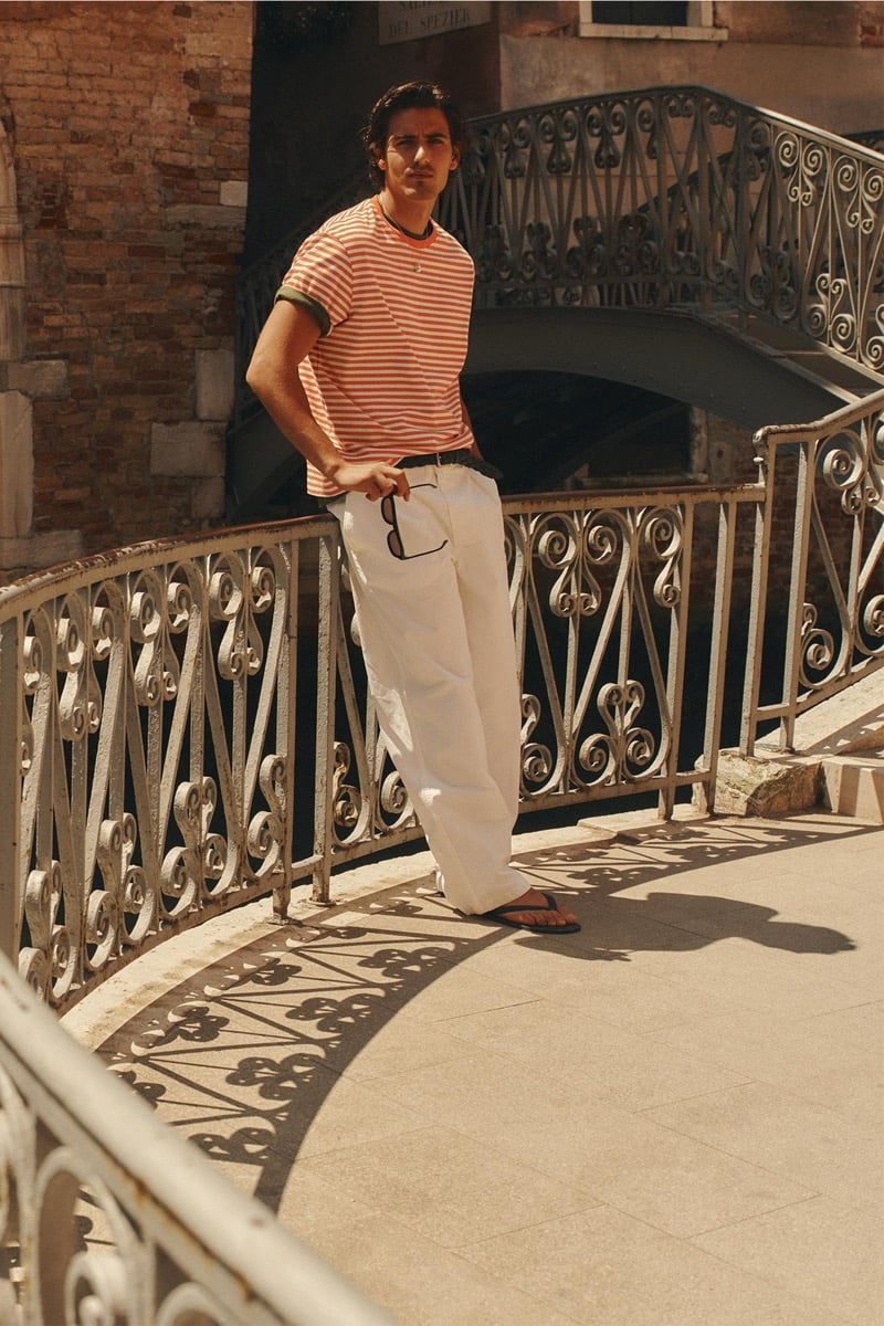 Model Tony Özkan in an orange and cream striped ringer tee with light wide-leg trousers, standing on an ornate wrought-iron Venetian footbridge holding sunglasses