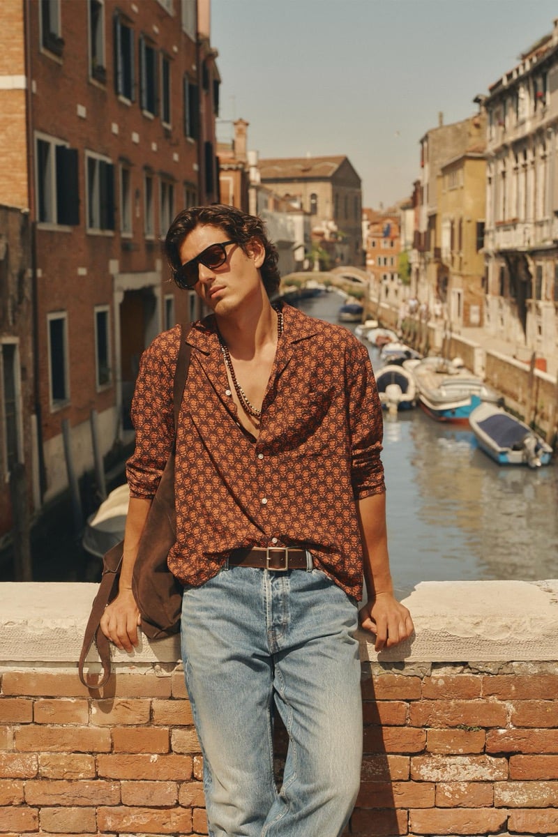 Model Tony Özkan in a rust-toned printed camp-collar shirt worn open over light blue faded jeans, sunglasses on and a brown suede shoulder bag, leaning against a brick wall above a Venice canal lined with boats