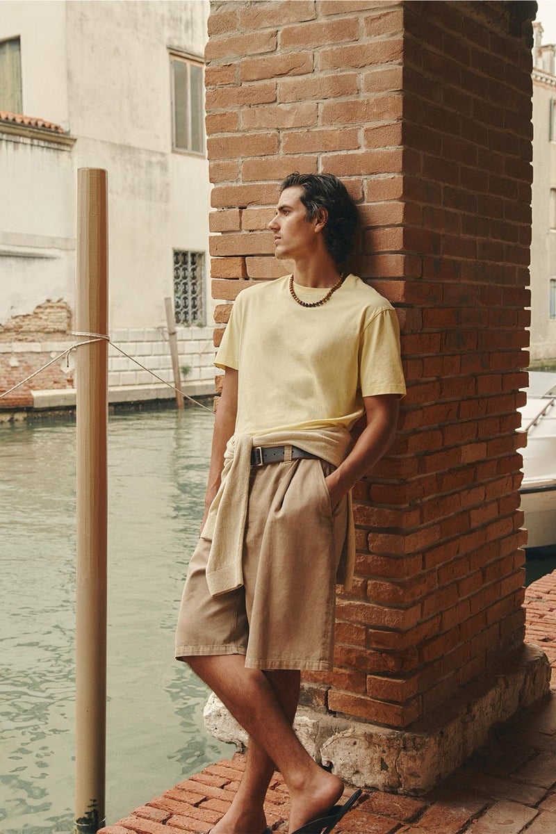 Model Tony Özkan in a soft yellow short-sleeve tee tucked into tan pleated shorts with a beige knit sweater tied at the waist, leaning against a brick column beside a canal