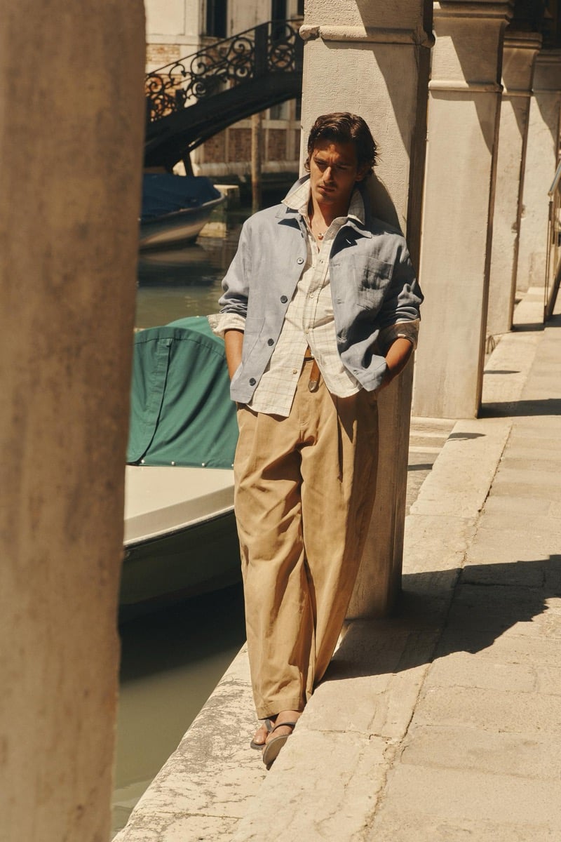 Model Tony Özkan leaning against a stone column beside a Venice canal in a blue linen chore jacket layered over a checked shirt with tan pleated wide-leg trousers and sandals