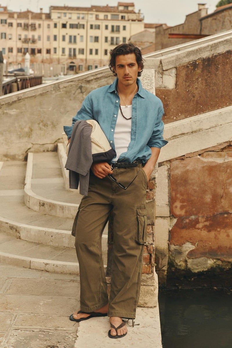 Model Tony Özkan in a faded denim shirt worn open over a ribbed tank, paired with olive cargo pants and flip-flops on weathered stone steps beside a canal