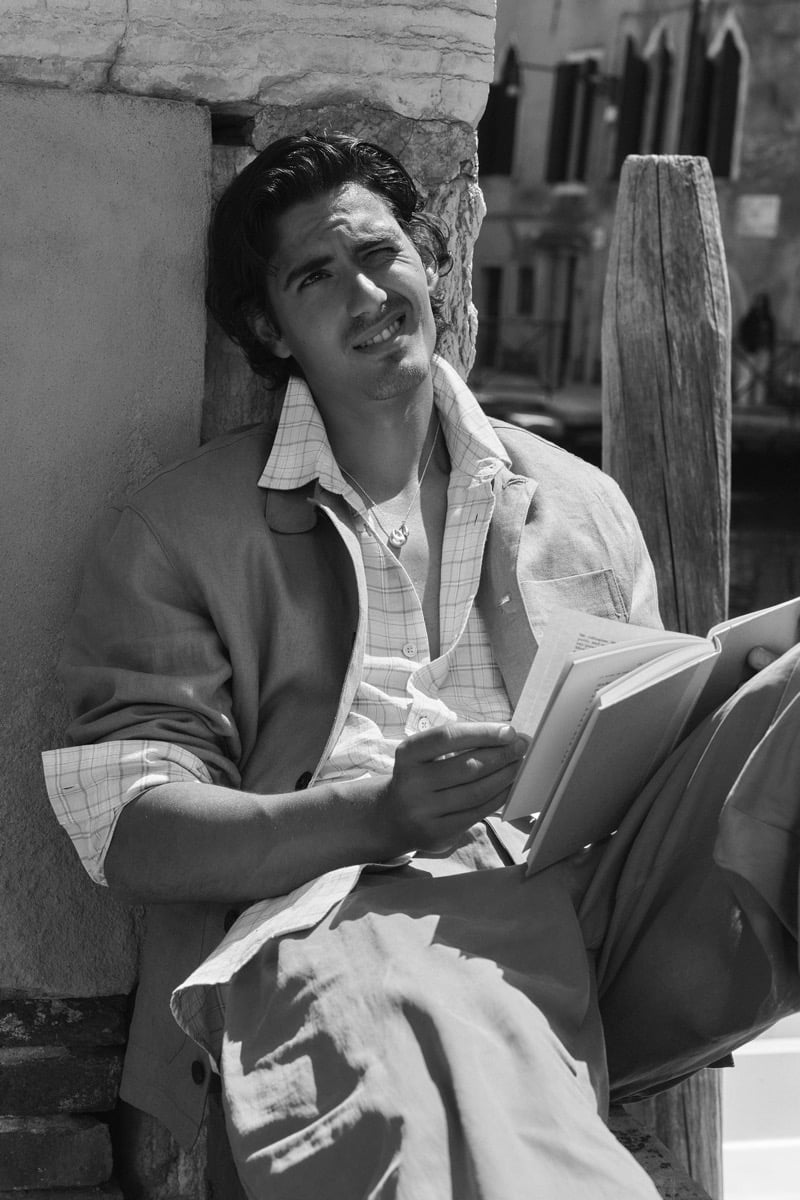Black-and-white portrait of model Tony Özkan reading a book outdoors in a beige linen overshirt and trousers layered over a checked shirt, seated against a brick wall